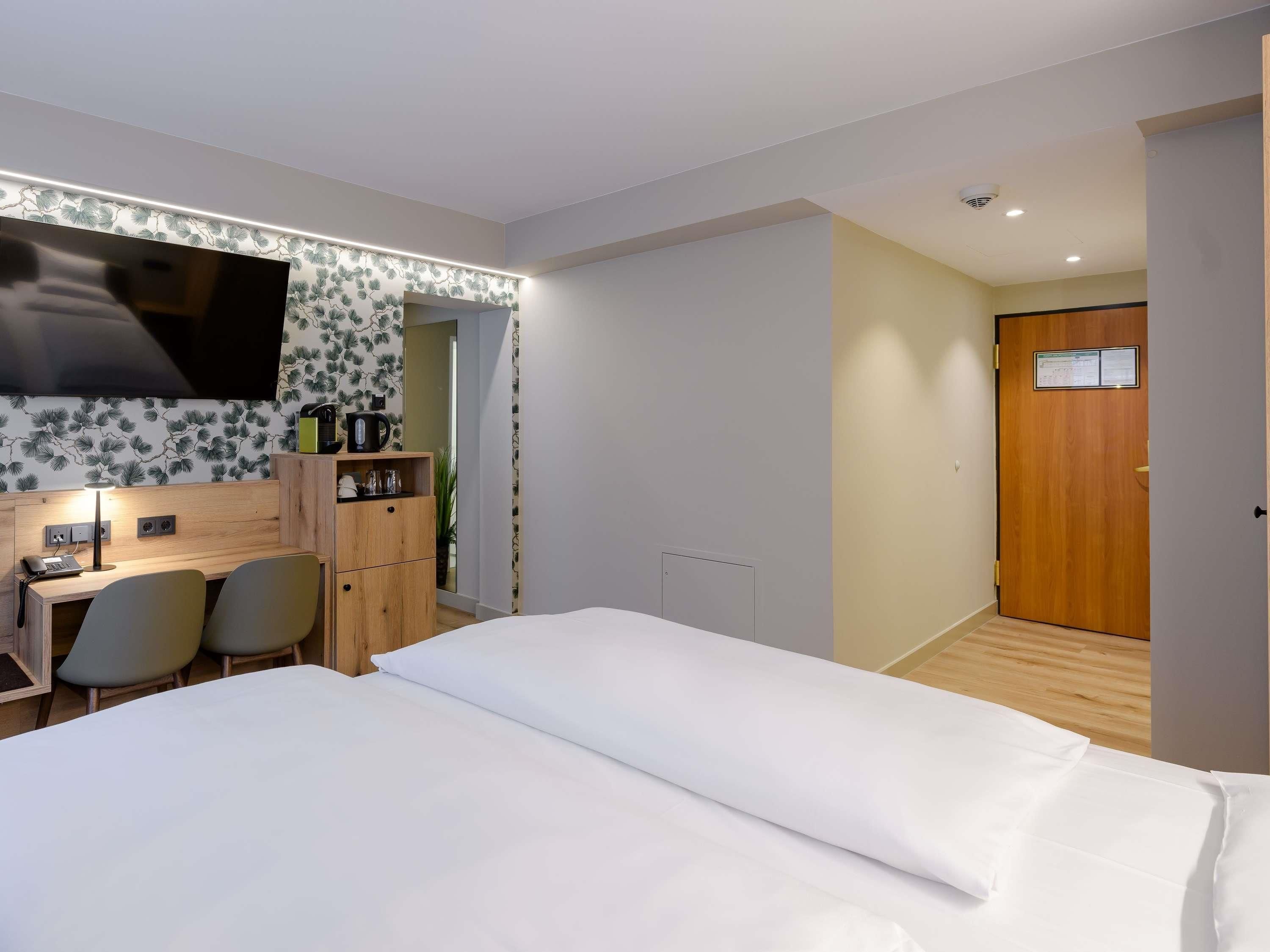 Una camera d'albergo con un letto, una scrivania con una sedia, una TV fissata al muro e una porta in legno.