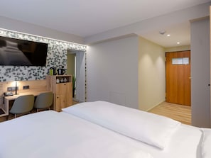 Habitación de hotel con cama, escritorio y silla, televisor montado en la pared y puerta de madera.