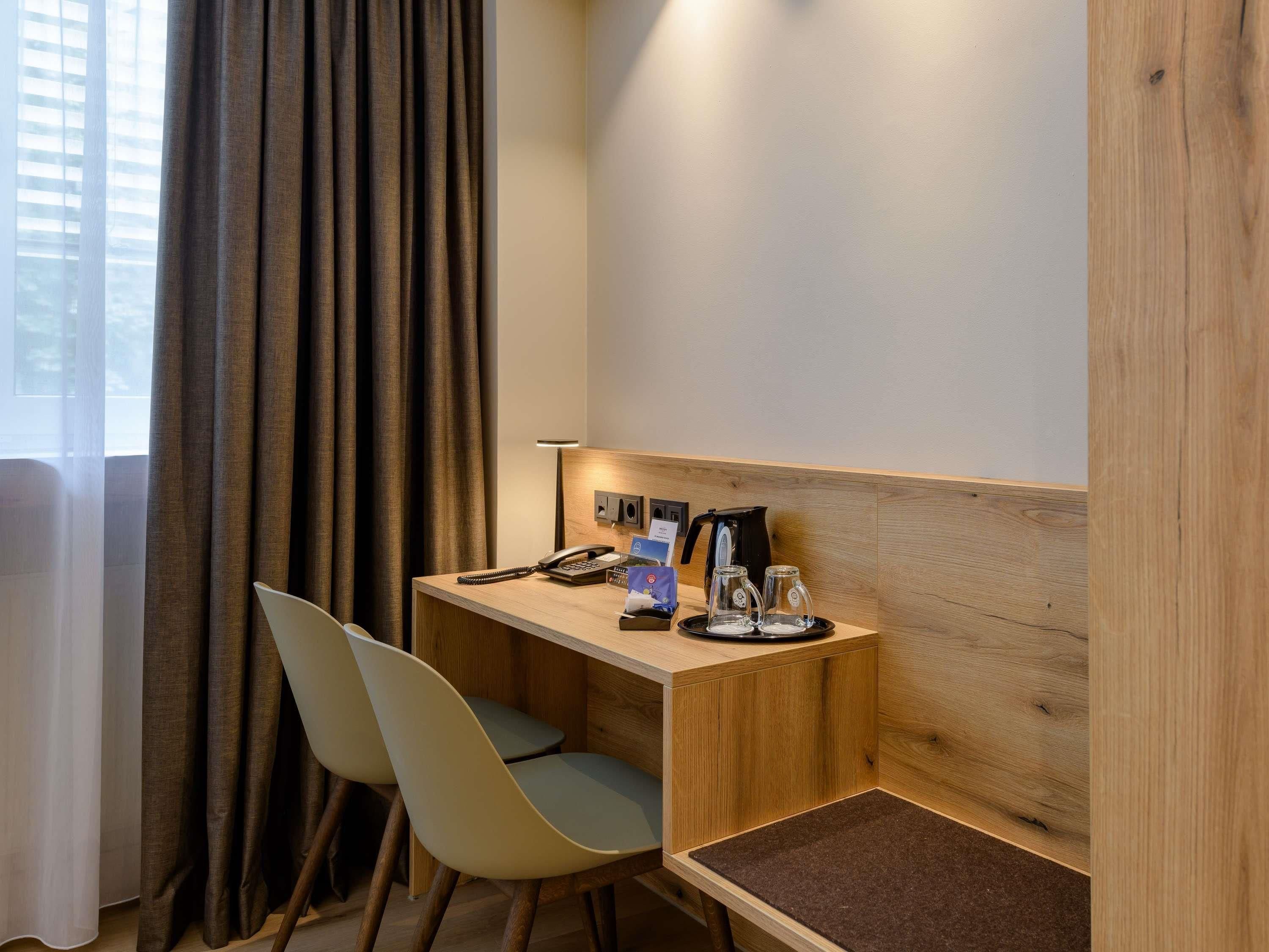 Una habitación de hotel con un escritorio de madera, una silla, un teléfono, una cafetera y una lámpara.