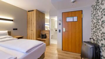 Habitación de hotel con cama, armario de madera, escritorio, maleta y baño visible a través de una puerta abierta.