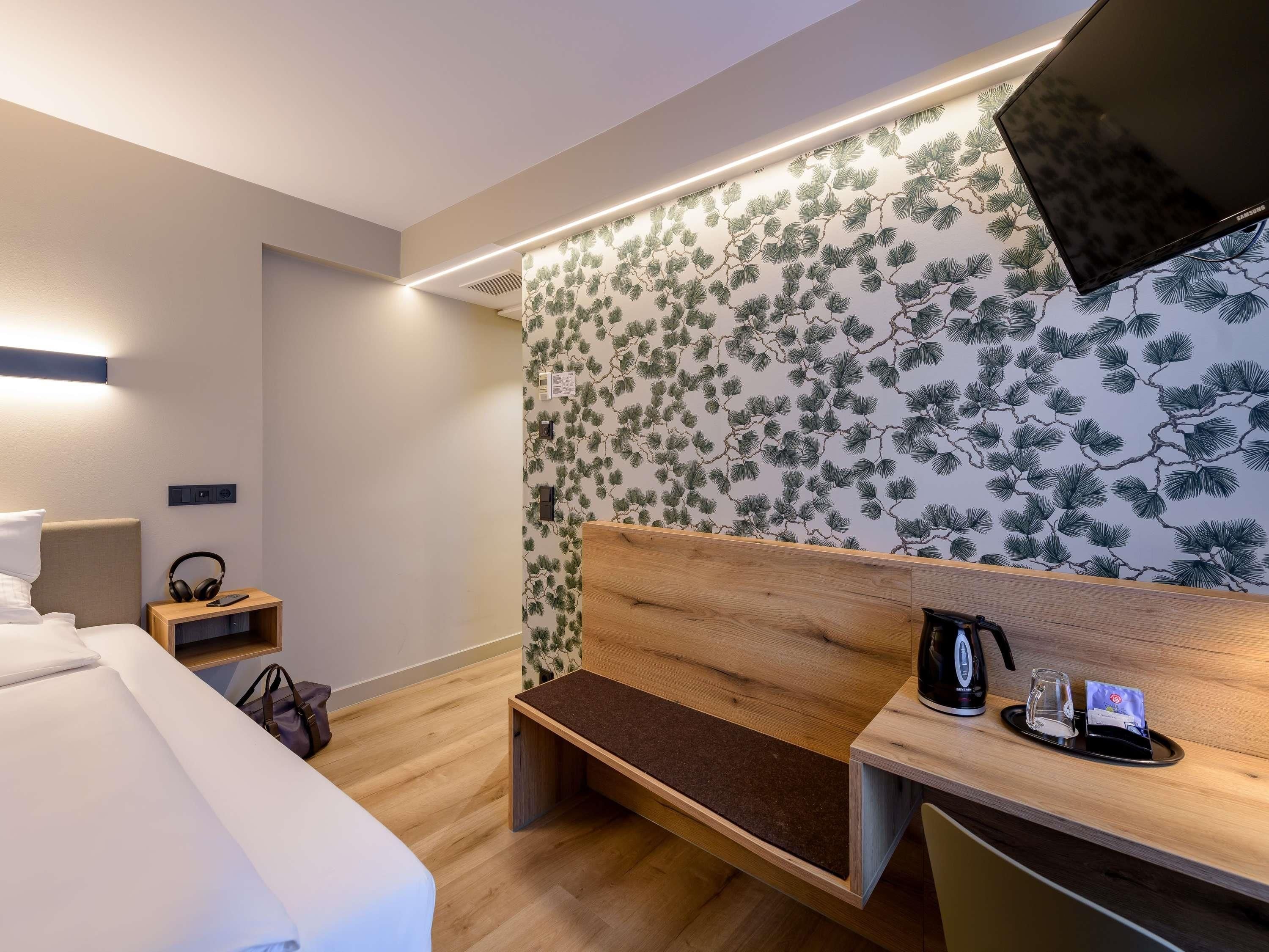 A modern hotel room with a bed, a wooden bench, a desk with a coffee maker, and a wall with a pine tree pattern.