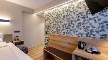 A modern hotel room with a bed, a wooden bench, a desk with a coffee maker, and a wall with a pine tree pattern.