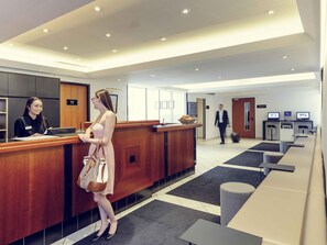 Een hotel receptie met een houten balie, een wachtruimte met banken en een digitaal informatiepunt.