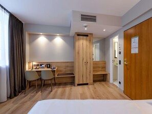 A hotel room with a bed, a desk with chairs, a wooden wardrobe, a mirror, and a door with a sign. - Mercure Hotel München Altstadt (Munich)