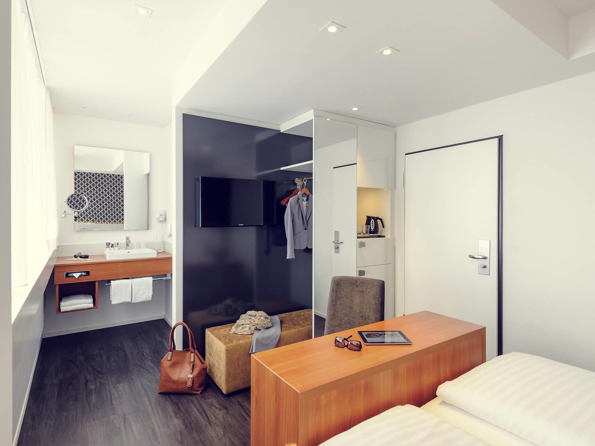 A modern hotel room with a wooden desk, a bed, a small seating area, a mirror, a TV, and a wardrobe.