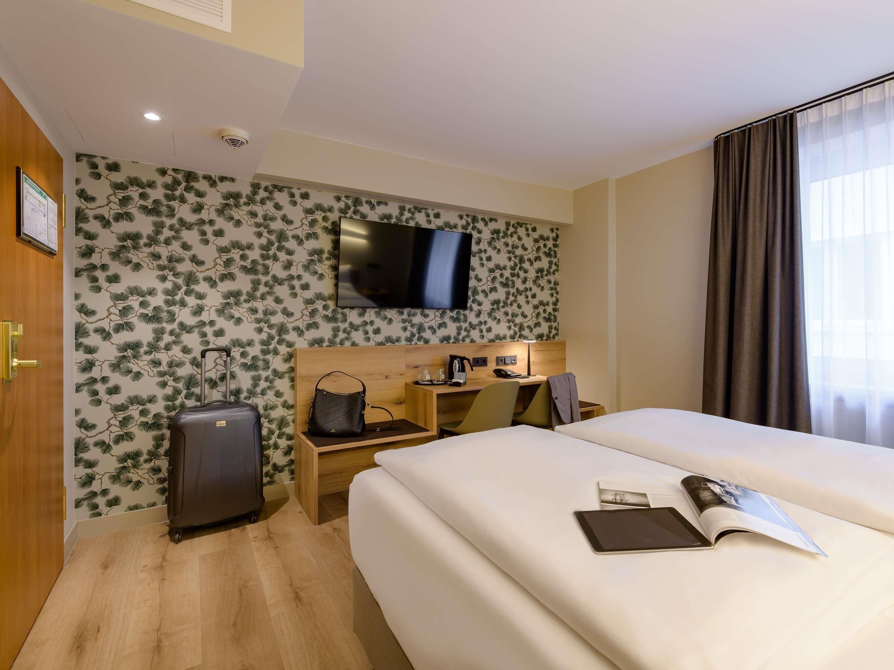 Habitació d'hotel amb un llit, una taula de treball amb una cadira, una televisió, una maleta i una finestra amb cortines.