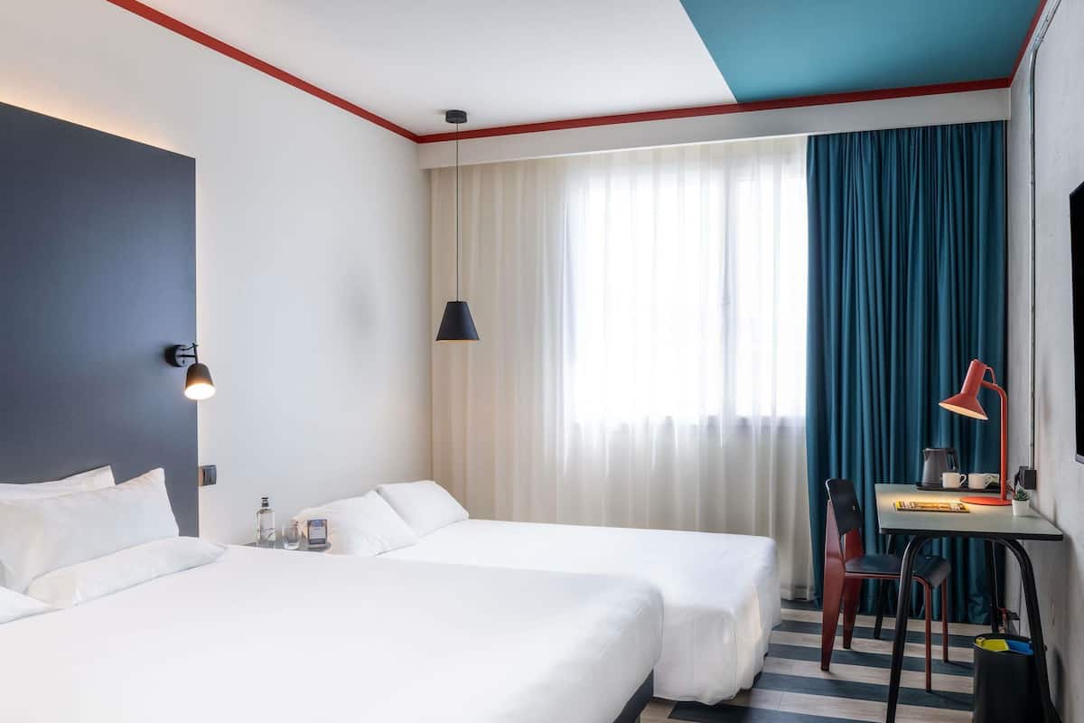 Une chambre d’hôtel avec deux lits, un bureau avec une lampe, une chaise, une petite table et une fenêtre avec des rideaux.