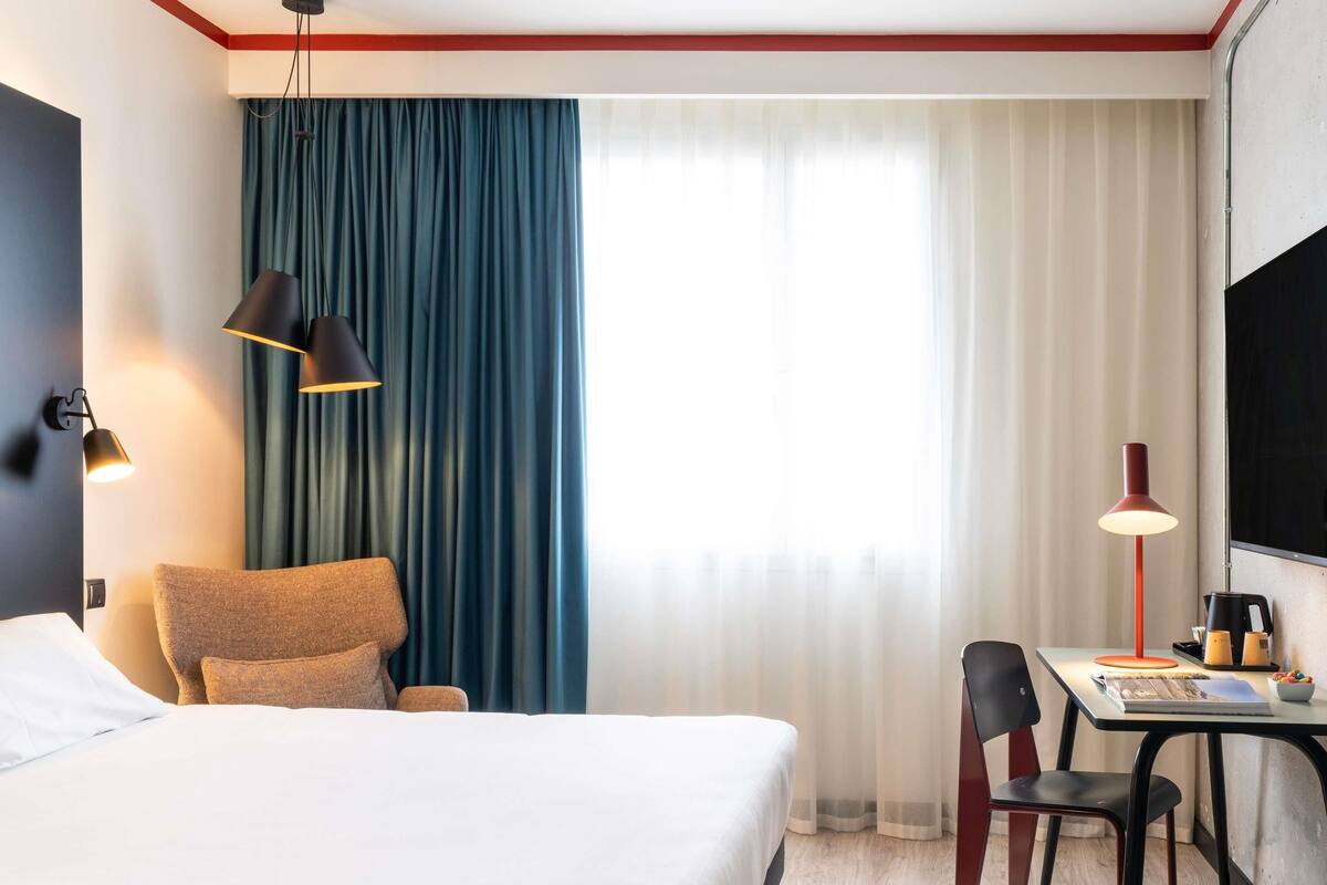 Une chambre d’hôtel avec un lit, un bureau, deux chaises, une télévision et une fenêtre avec des rideaux.