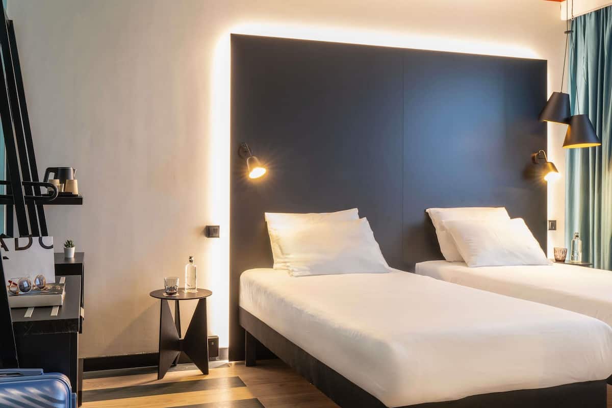 Une chambre d’hôtel avec deux lits, une petite table de chevet, une lampe et une étagère avec quelques objets.