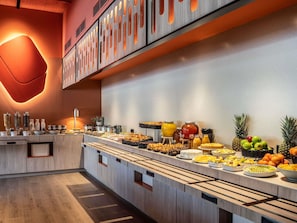 Un bufé con diversos alimentos y bebidas, que incluye frutas y pasteles, en un ambiente moderno con paneles de madera y una lámpara que emite luz cálida.