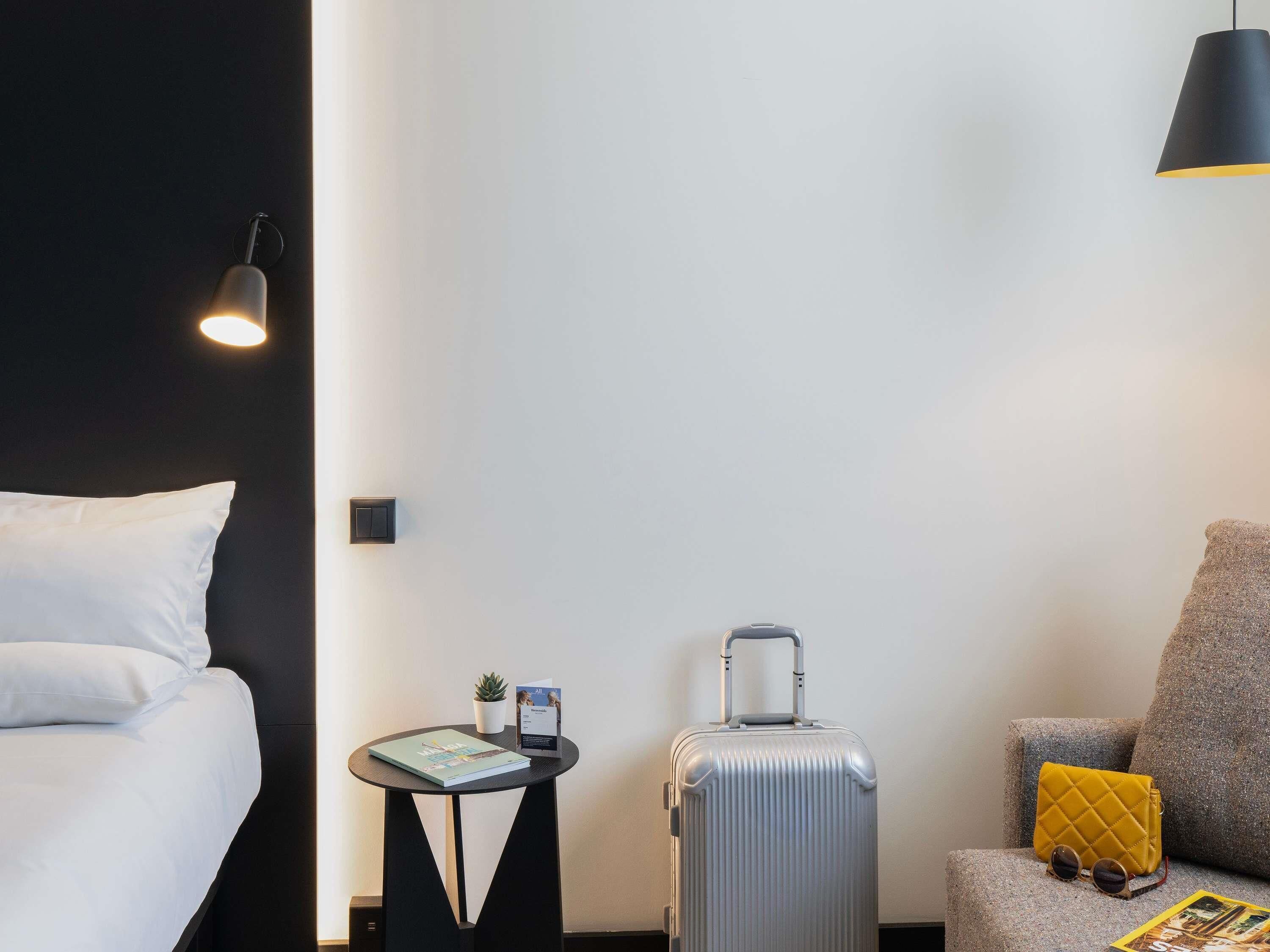 Habitación de hotel con una cama, una mesita con un libro y una planta, una maleta y una silla con cojín.