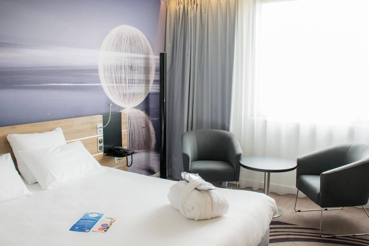 Hotellihuone, jossa on sänky, kaksi tuolia, pieni pöytä, televisio ja näkymä merelle.
