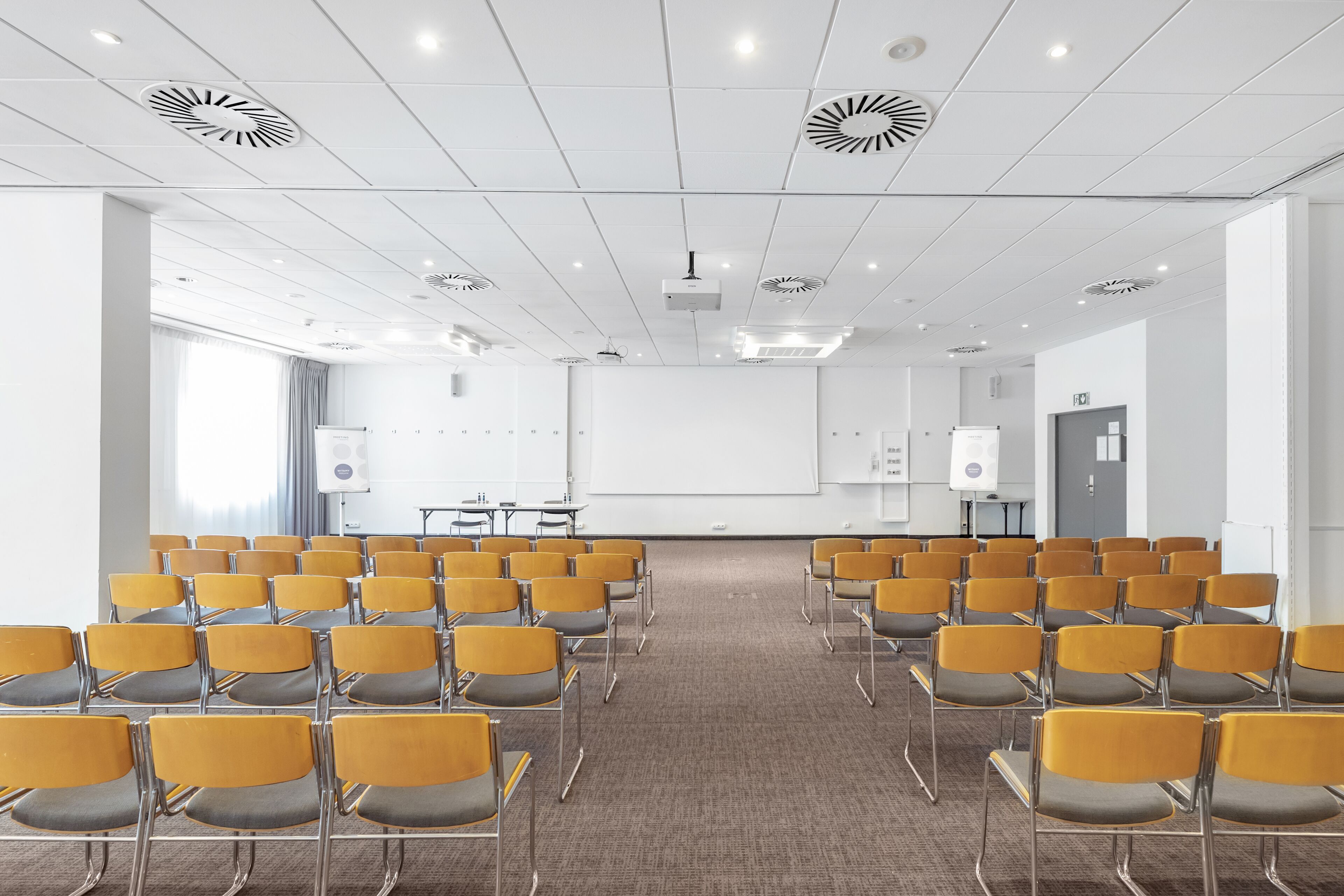A conference room with rows of yellow chairs, a whiteboard, and a projector on the ceiling.