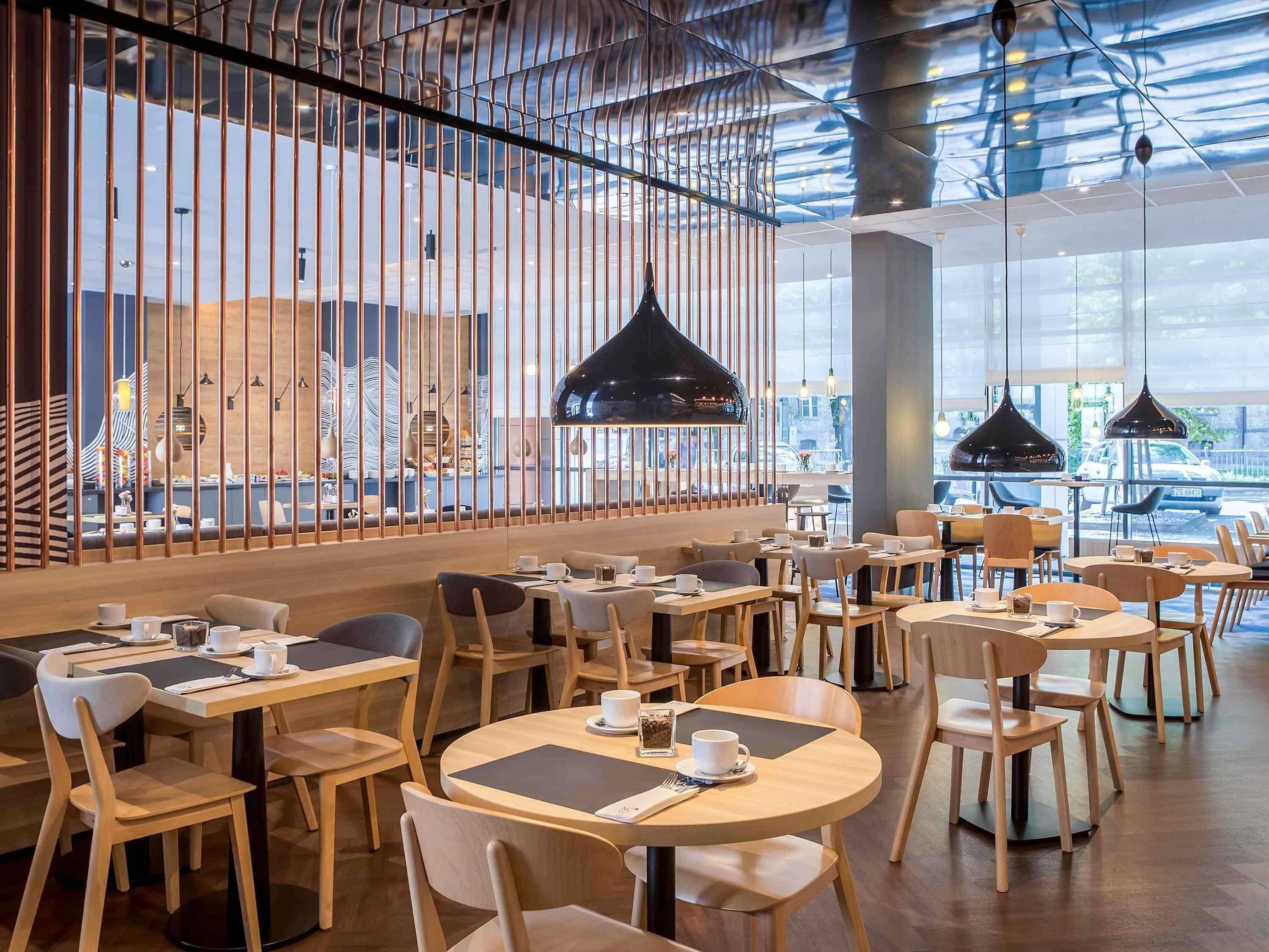 A modern cafe interior with wooden tables and chairs, black pendant lights, and a view through large windows.