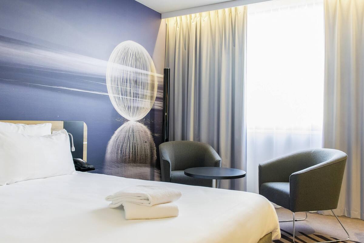 Hotellihuone, jossa on sänky, kaksi tuolia, pieni pöytä, seinätaideteos ja ikkuna, jossa on verhot.