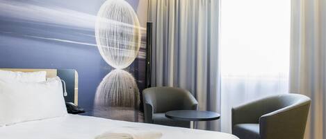 A hotel room with a bed, two chairs, a small table, a wall art piece, and a window with curtains.