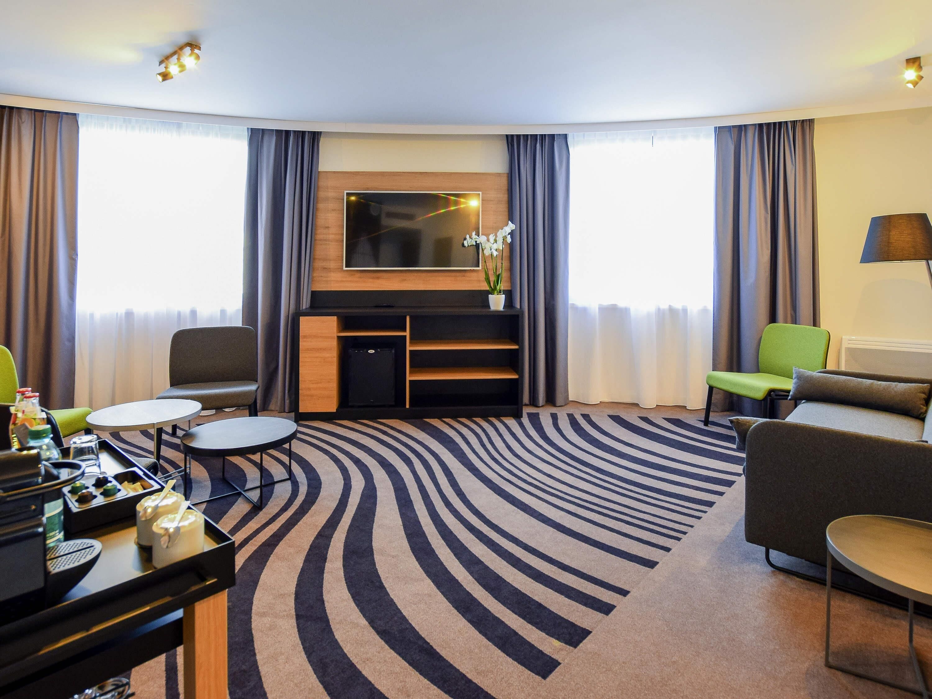 A hotel room with a curved carpet, a flat-screen TV, a sofa, a chair, a small table, and a minibar.