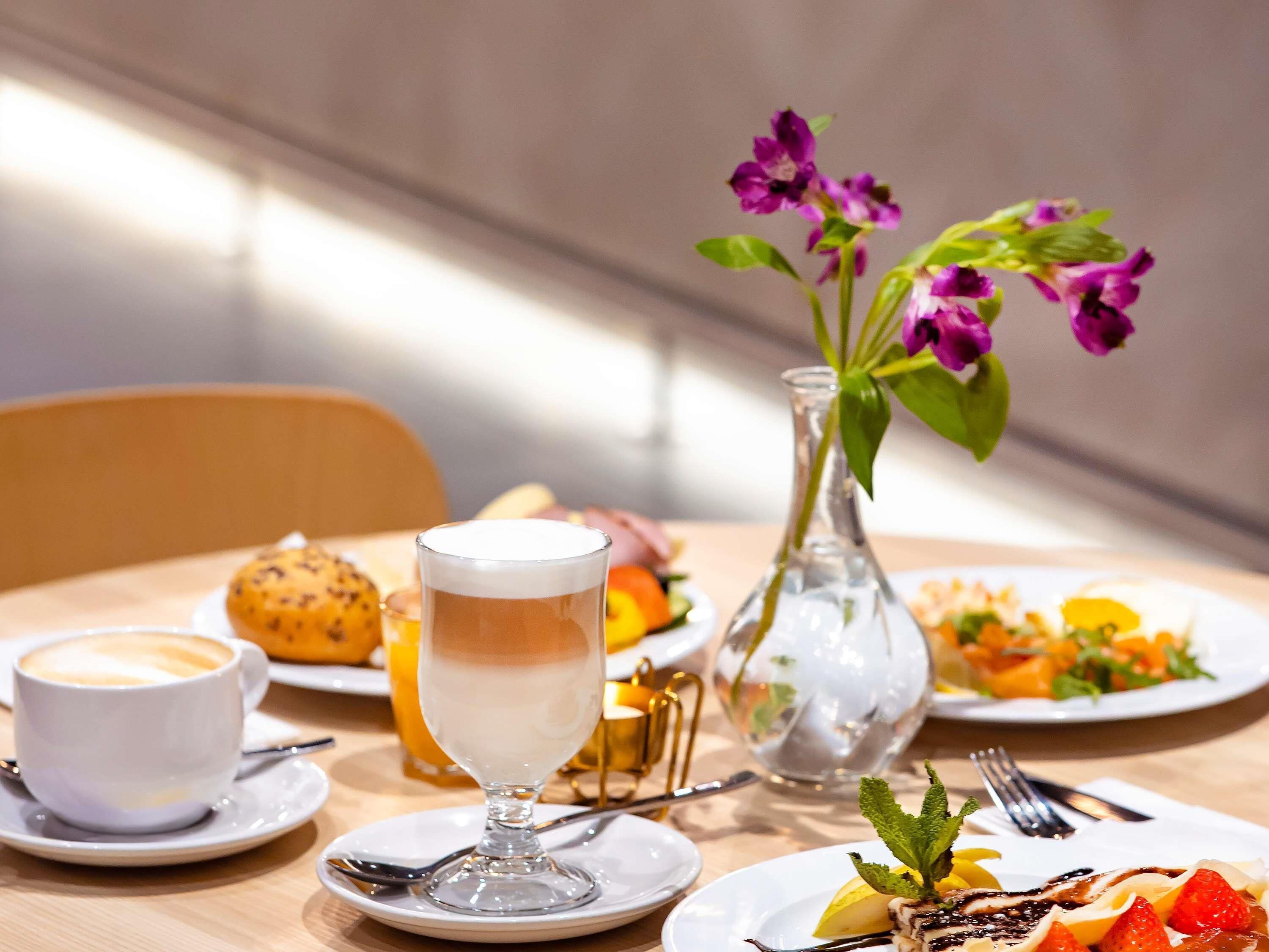 Una mesa con una taza de café, un vaso de leche, un plato de comida, un florero con flores y un frutero.