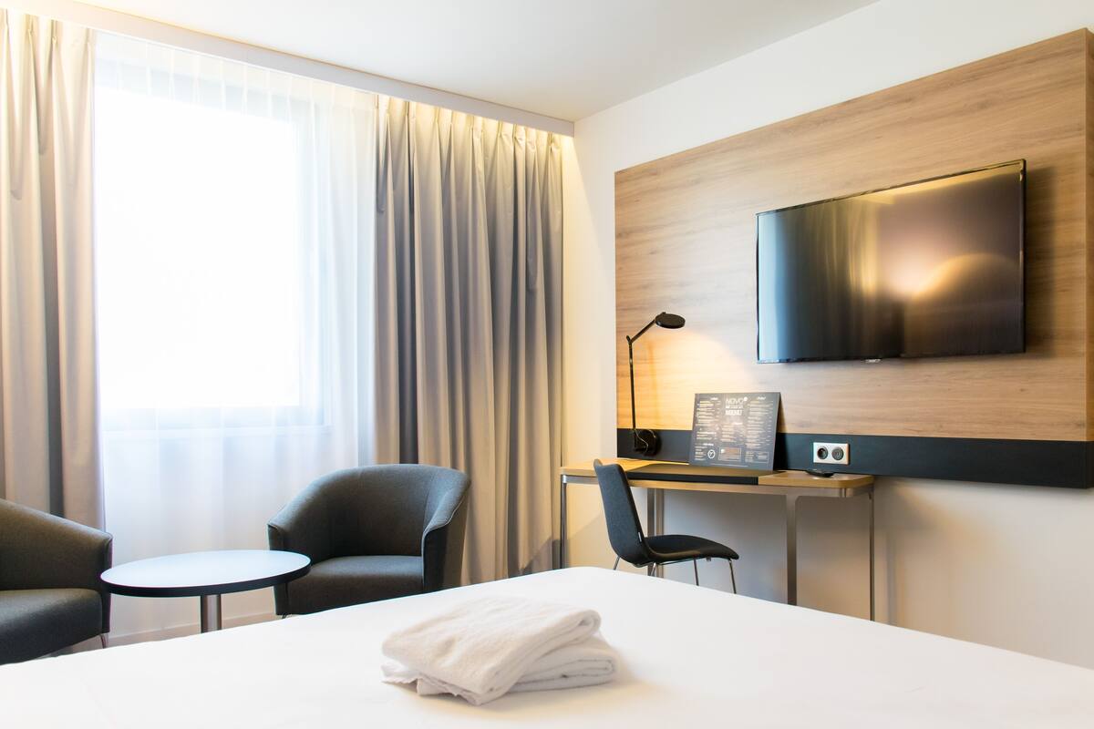Hotellihuone, jossa on sänky, työpöytä tietokoneella, kaksi tuolia, seinälle asennettu televisio ja ikkuna, jossa on verhot.