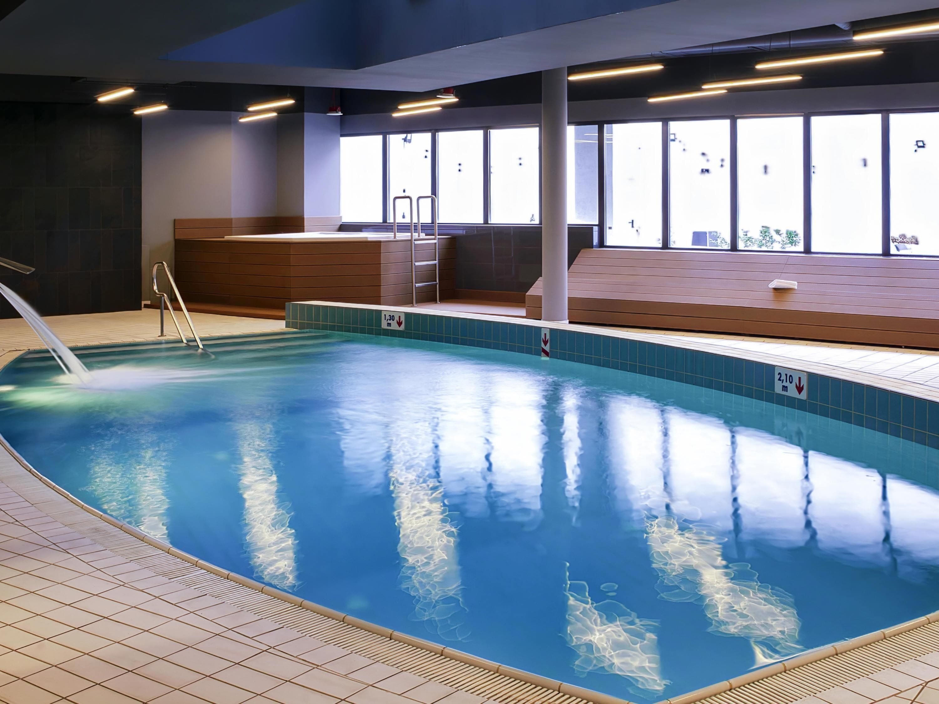 Una piscina interior con agua cristalina, suelo de baldosas y una zona con bancos de madera.