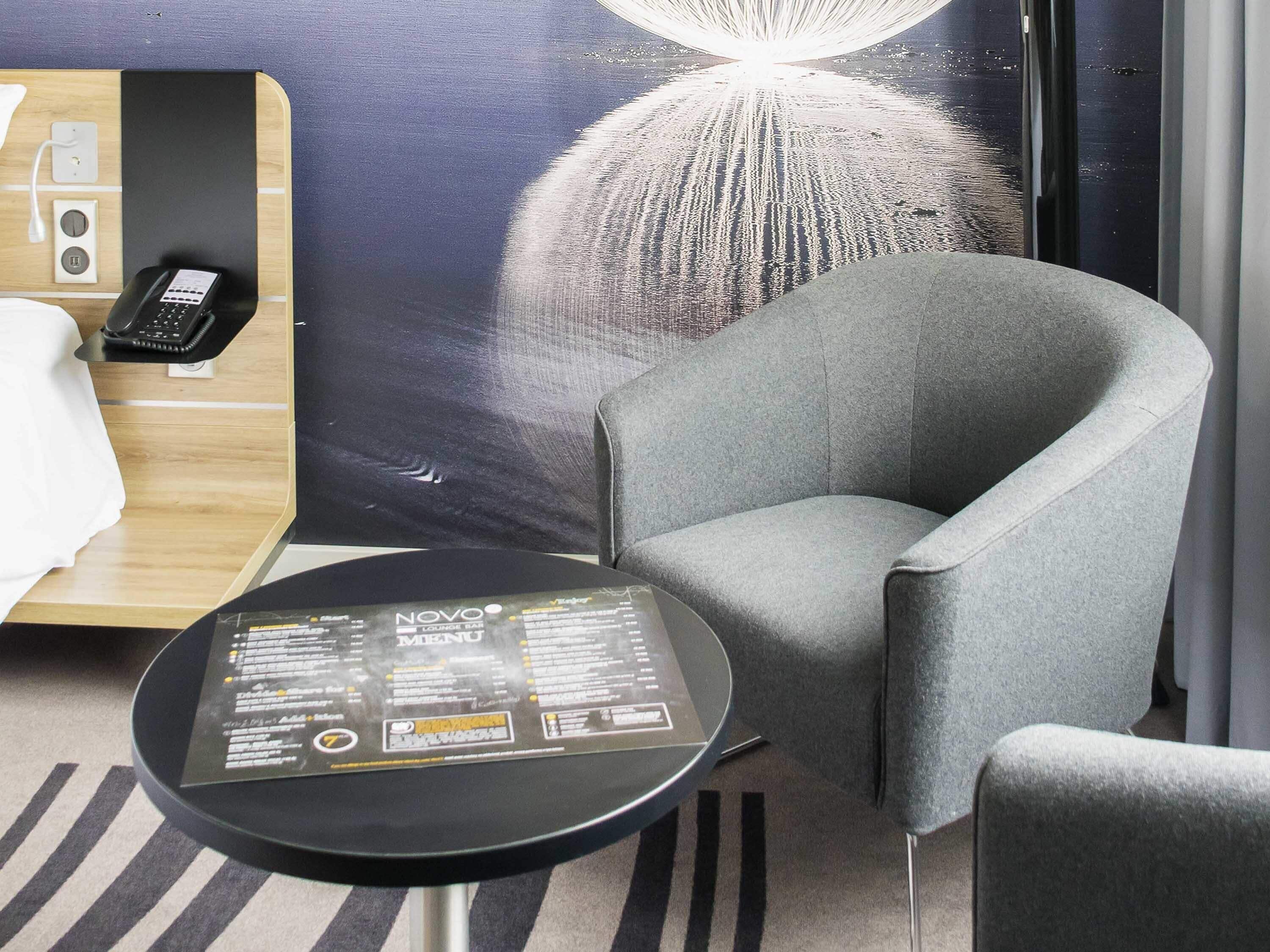 Une chambre d’hôtel avec un fauteuil gris, une petite table ronde sur laquelle se trouve un menu, un lit et un téléphone fixé au mur.