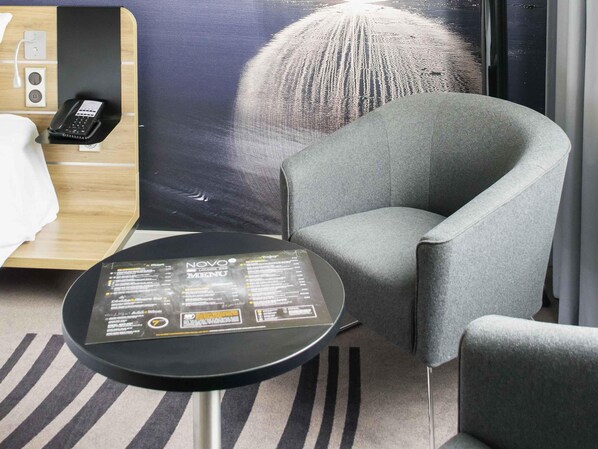 Une chambre d’hôtel moderne avec un fauteuil gris, une petite table ronde sur laquelle est posé un menu, et une grande affiche représentant une structure sphérique.