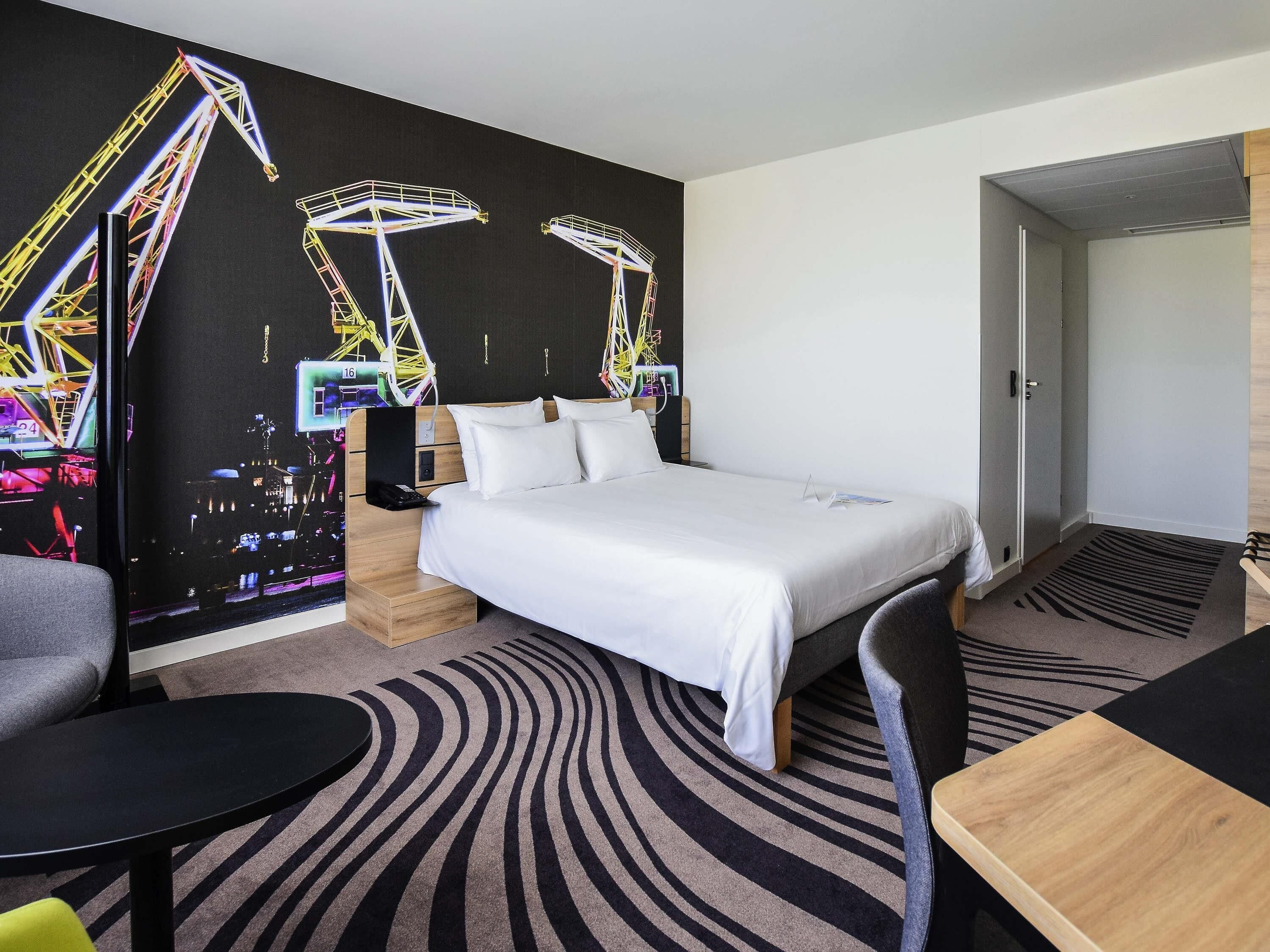 A hotel room with a bed, a desk, a chair, a small table, and a wall with a colorful mural.