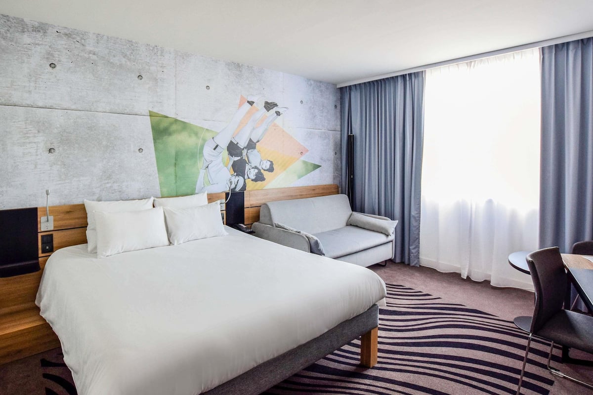 A hotel room with a bed, a sofa, a chair, a small table, and a painting on the wall.