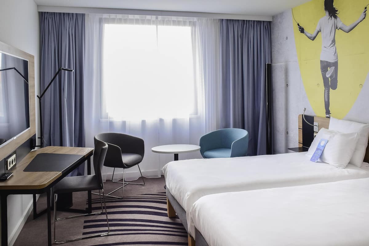 A hotel room with a bed, a desk, a chair, a small table, and a large window with curtains.