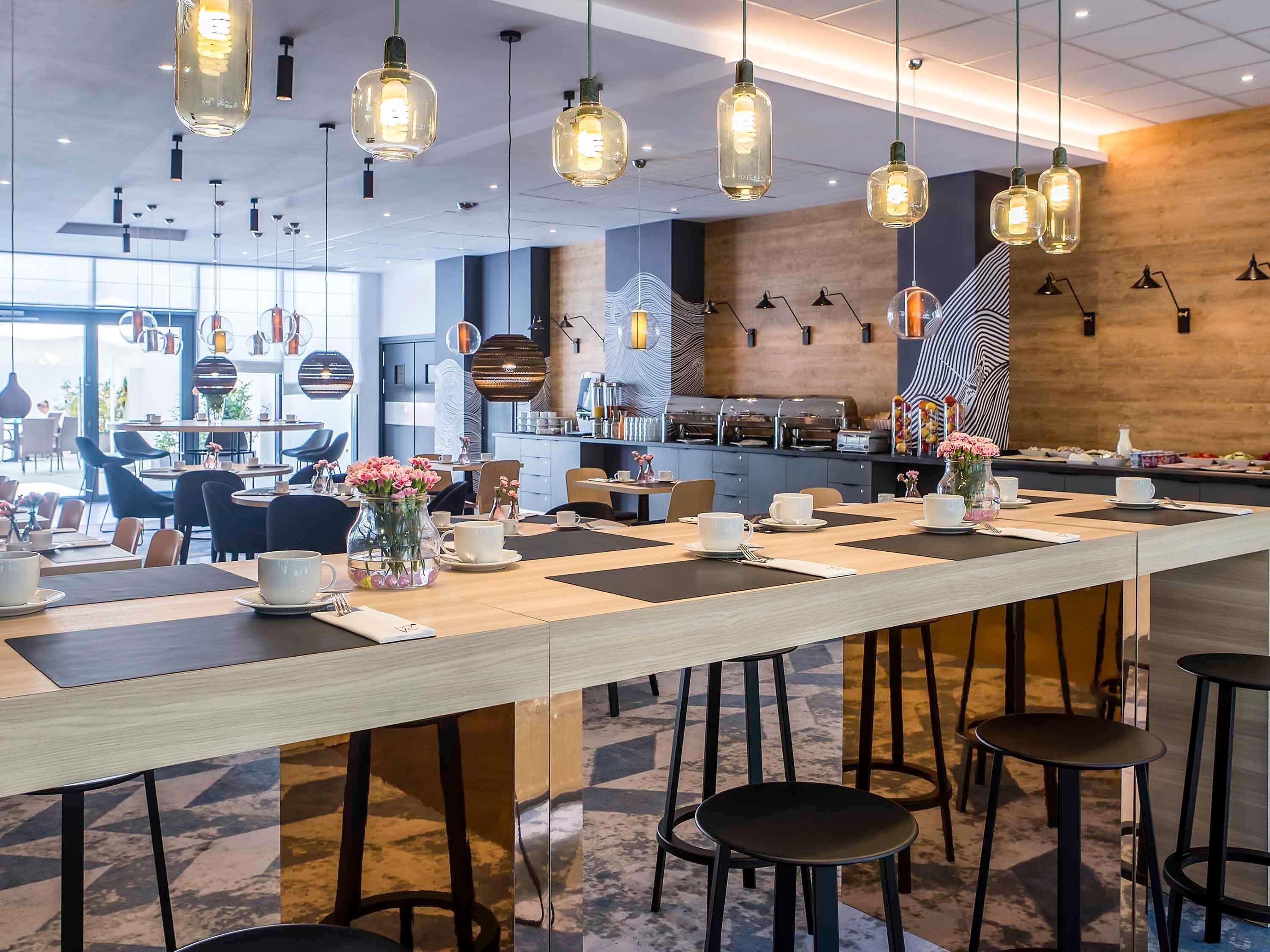 A modern cafe with a long communal table, hanging pendant lights, and a kitchen area in the background.