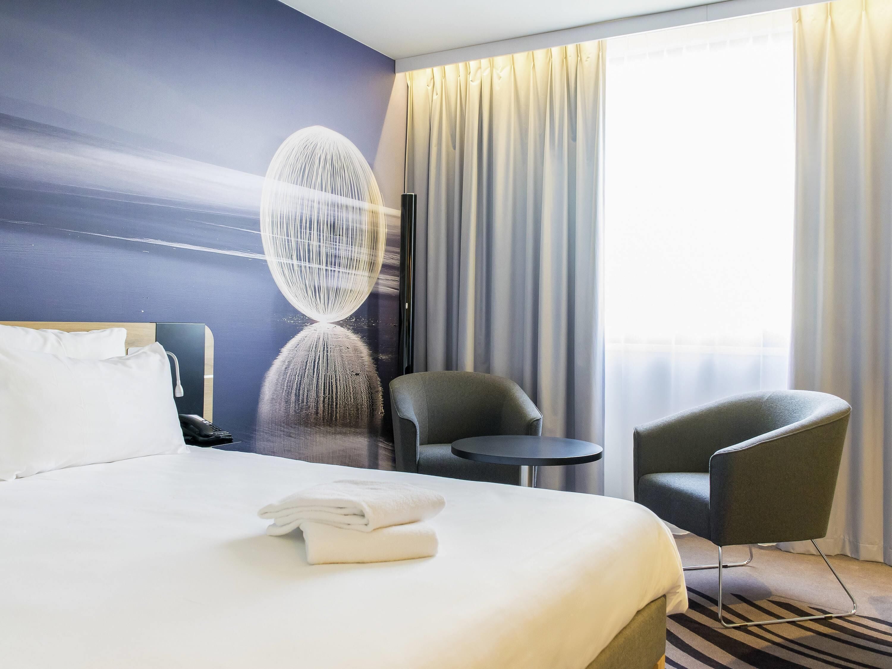 A hotel room with a bed, two chairs, a small table, a wall art piece, and a window with curtains.