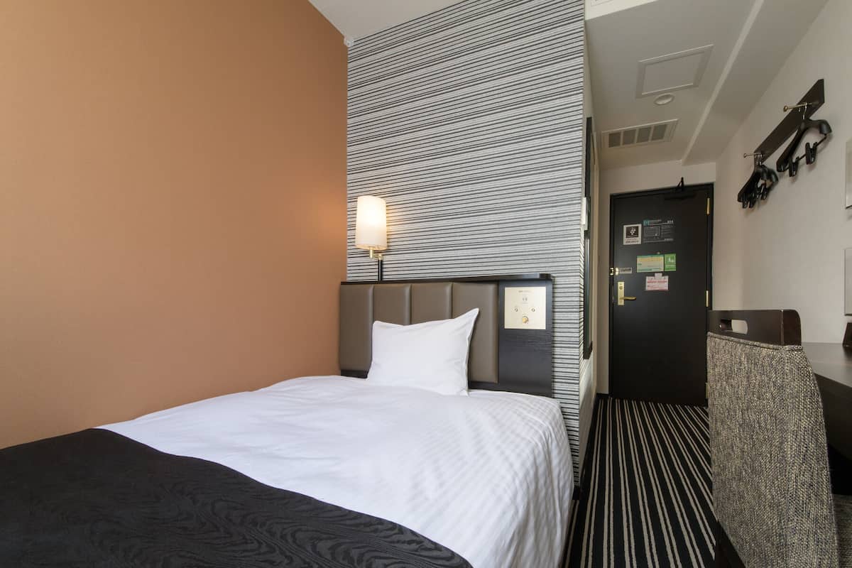 A hotel room with a bed, a nightstand, a wall-mounted lamp, a door with a keycard reader, and a coat rack.