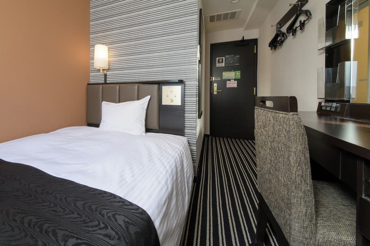 A hotel room with a bed, a desk, a chair, a wall-mounted lamp, and a door with a security camera.