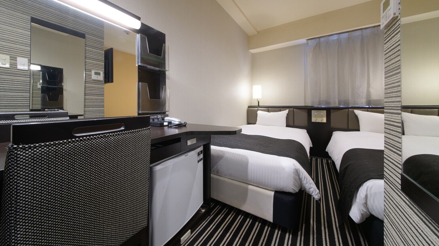 A hotel room with two beds, a desk, a small refrigerator, a microwave, and a large mirror.