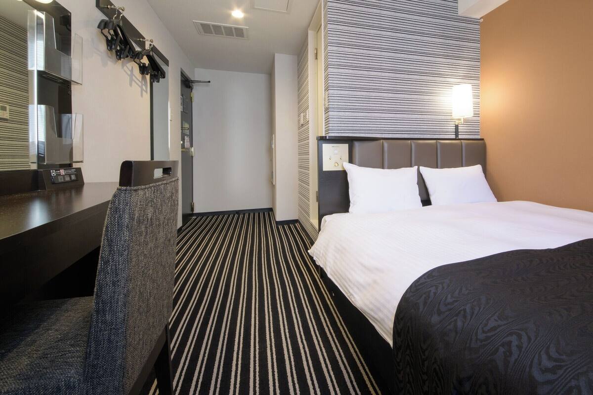 A hotel room with a bed, a desk, a chair, a mirror, and a wall-mounted control panel.