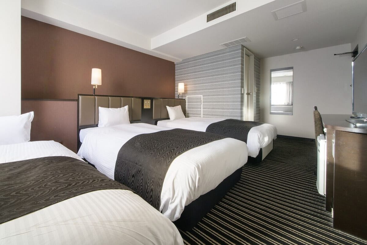 Ein Hotelzimmer mit drei Betten, einem Schreibtisch und einem großen Fenster.