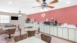 Une cafétéria avec des tables et des chaises, un coin café et un mur rouge orné d’un logo.