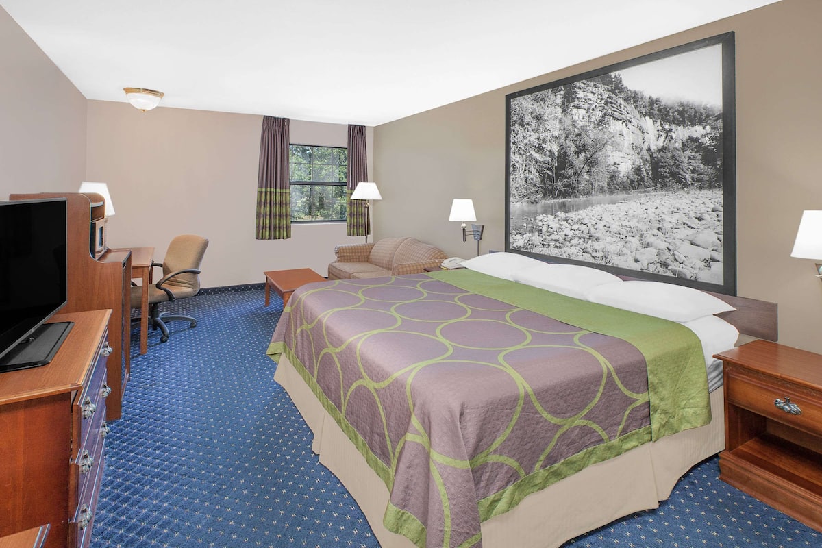 Ein Hotelzimmer mit einem großen Bett, einem Schreibtisch mit Fernseher, zwei Stühlen, einem Vorhangfenster und einem Bild an der Wand.