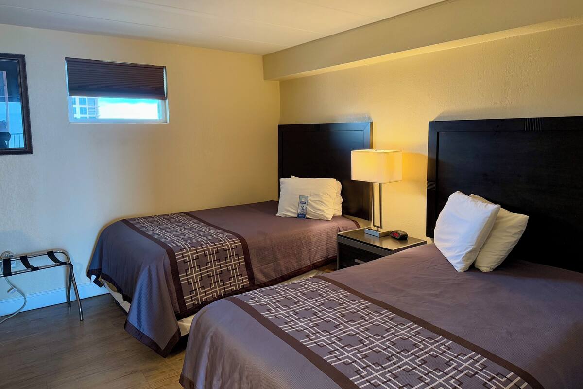 A hotel room with two beds, a nightstand with a lamp, a small table, and a window with blinds.