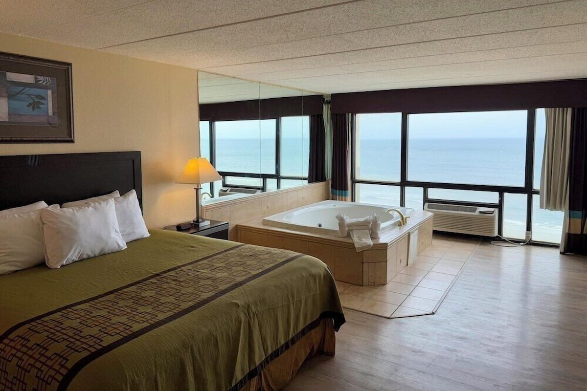 A hotel room with a large bed, a separate sitting area with a bathtub, a large window with a view of the sea, and a framed picture on the wall.