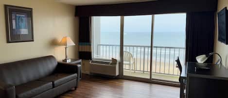 Habitación de hotel con sofá de cuero marrón oscuro, una mesita de noche con lámpara, un ventanal con vista a la playa y el océano, y un escritorio con silla.