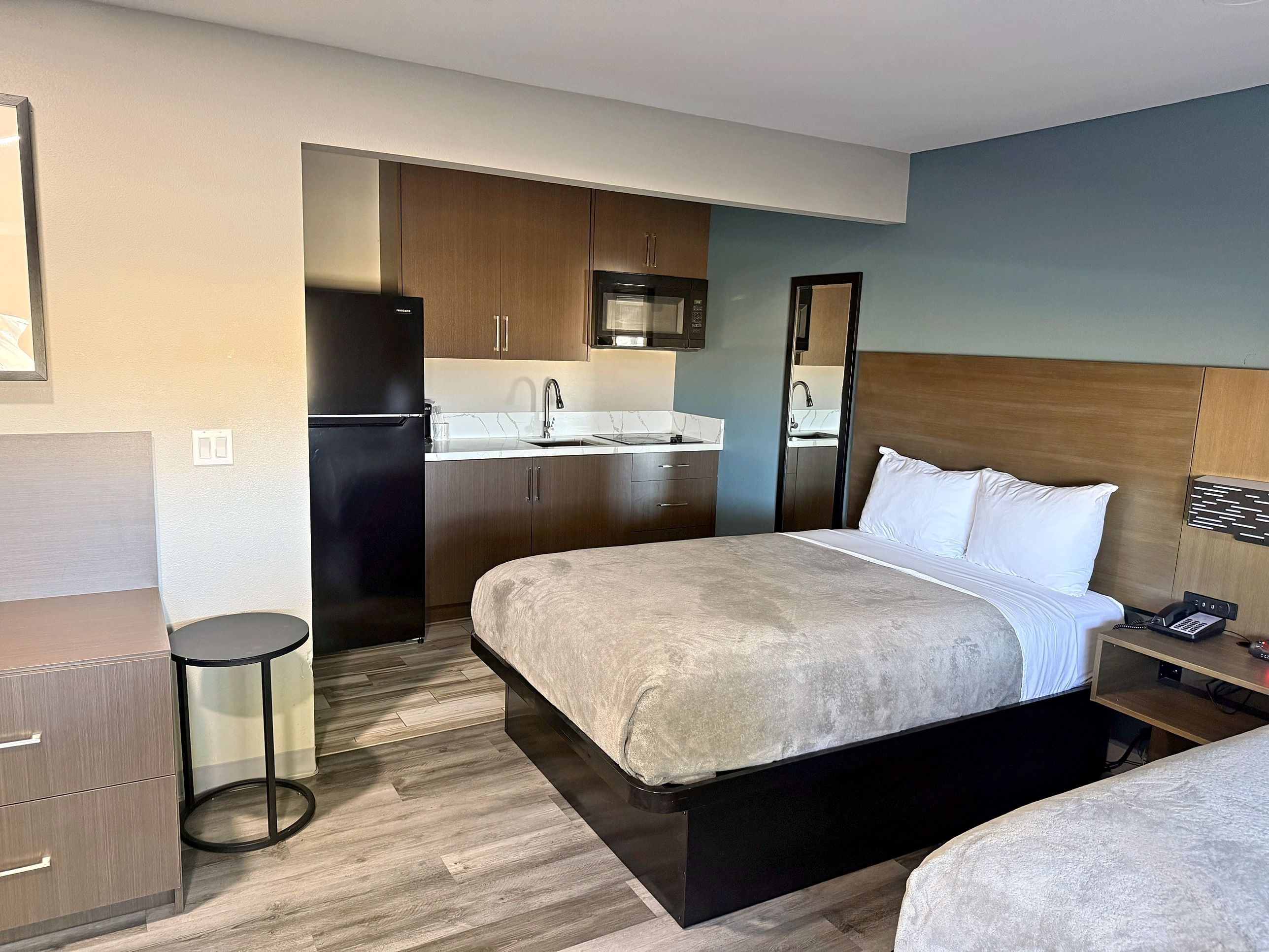 A hotel room with a bed, a desk, a chair, a small stool, a kitchenette, and a bathroom.