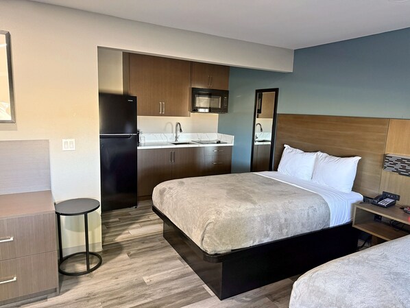 A hotel room with a bed, a desk, a chair, a small stool, a kitchenette, and a bathroom.