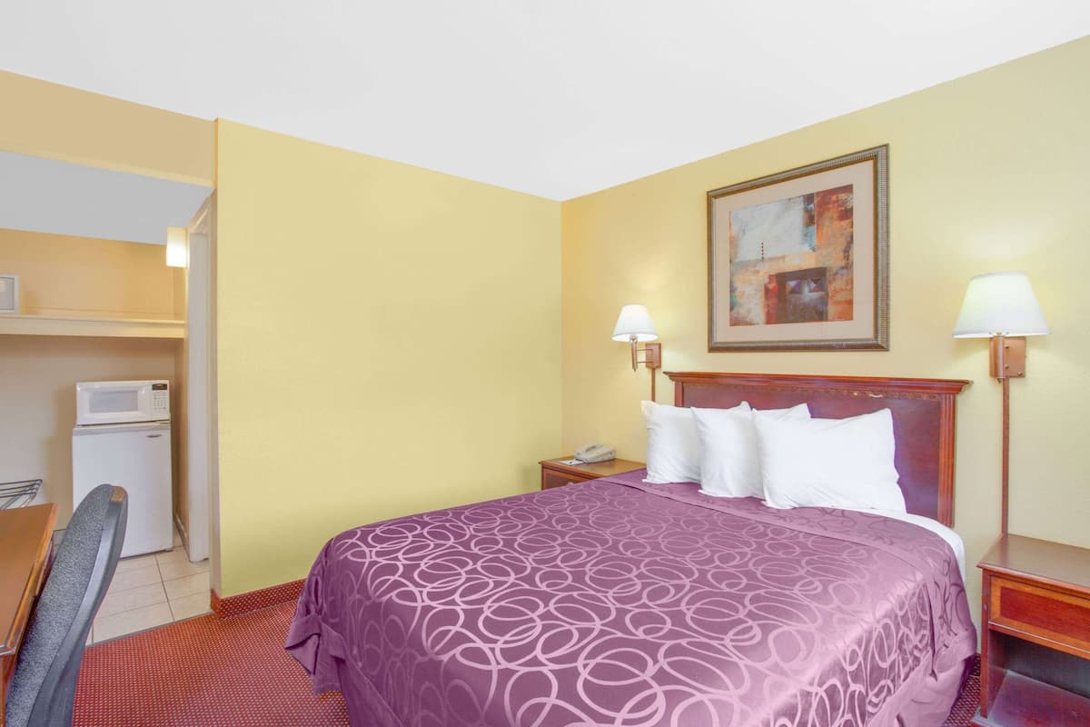 A hotel room with a bed, a desk, a chair, a microwave, a nightstand, and a painting on the wall.