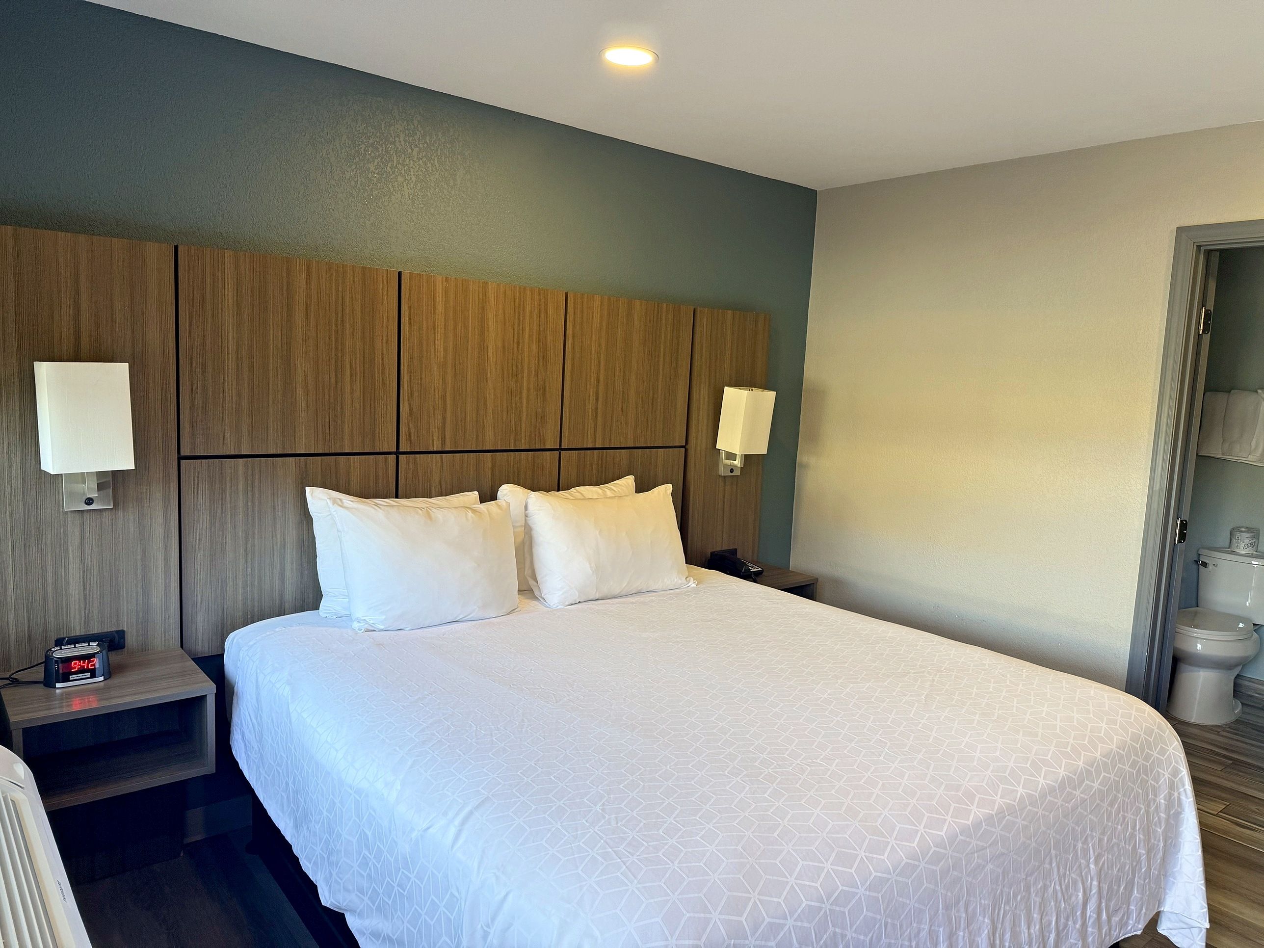 A hotel room with a large bed, two bedside tables, a nightstand, a wall-mounted lamp, and a visible clock.