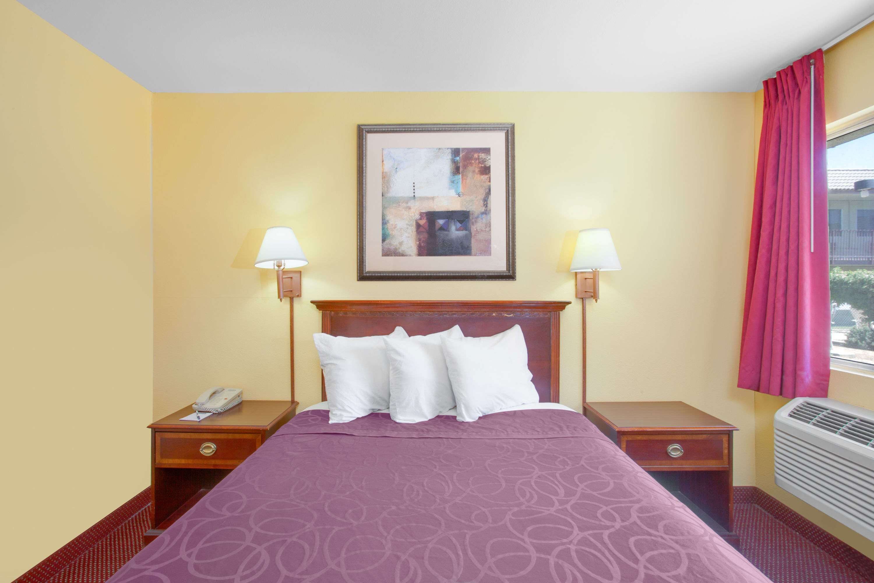 A hotel room with a bed, two bedside tables, a nightstand, a framed artwork, and a window with curtains.