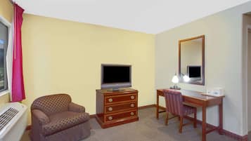 Una habitación de hotel con un sofá, un aparador de madera con cajones, un televisor, un escritorio con una silla, un espejo y una ventana con cortinas.