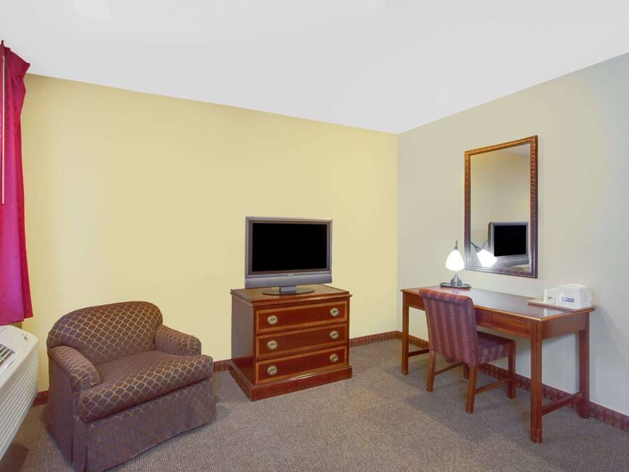 Una habitación de hotel con un sofá, un aparador de madera con cajones, un televisor, un escritorio con una silla, un espejo y una ventana con cortinas.