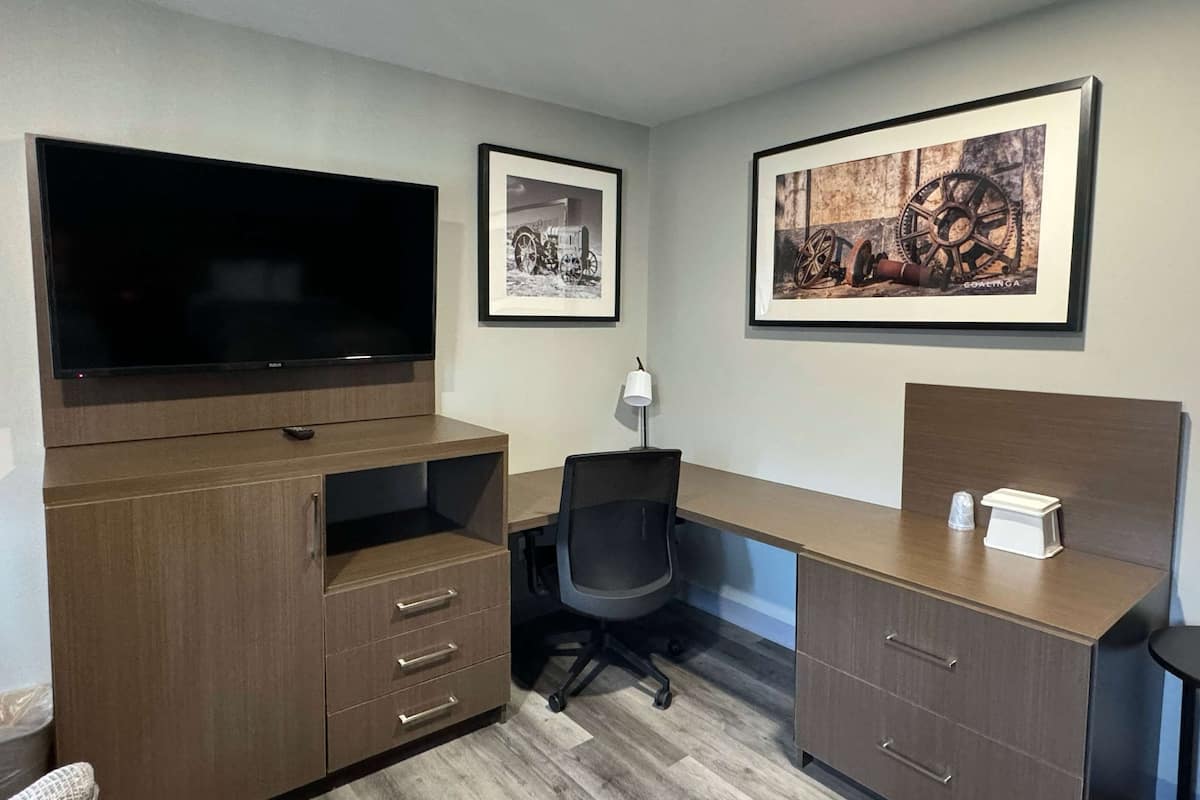 A modern office space with a large flat-screen TV, wooden desk, and office chair. There are framed pictures on the walls and a lamp on the desk.