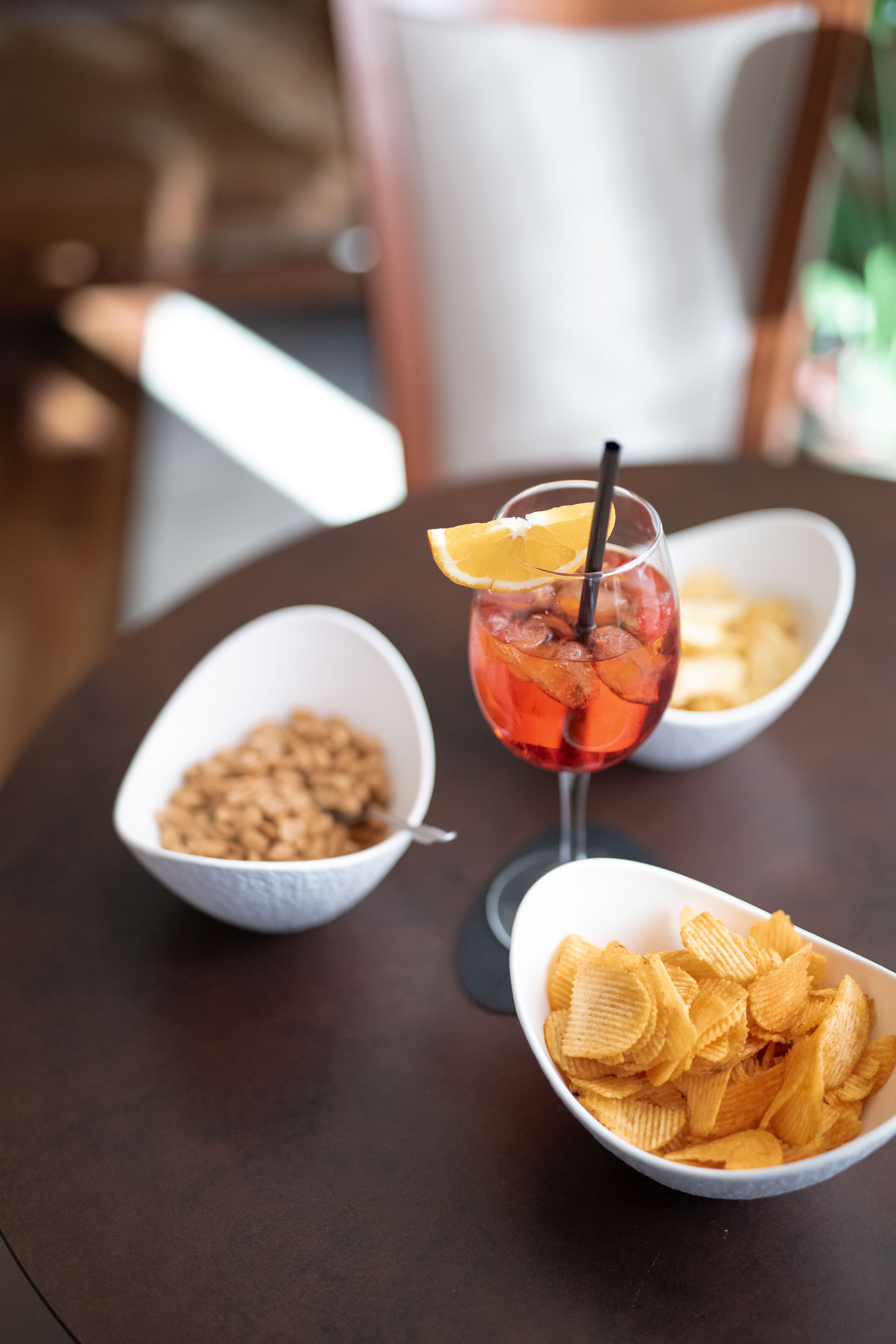 A table with three bowls containing different snacks, a glass of iced drink with a straw and a slice of orange, and a spoon in one of the bowls.